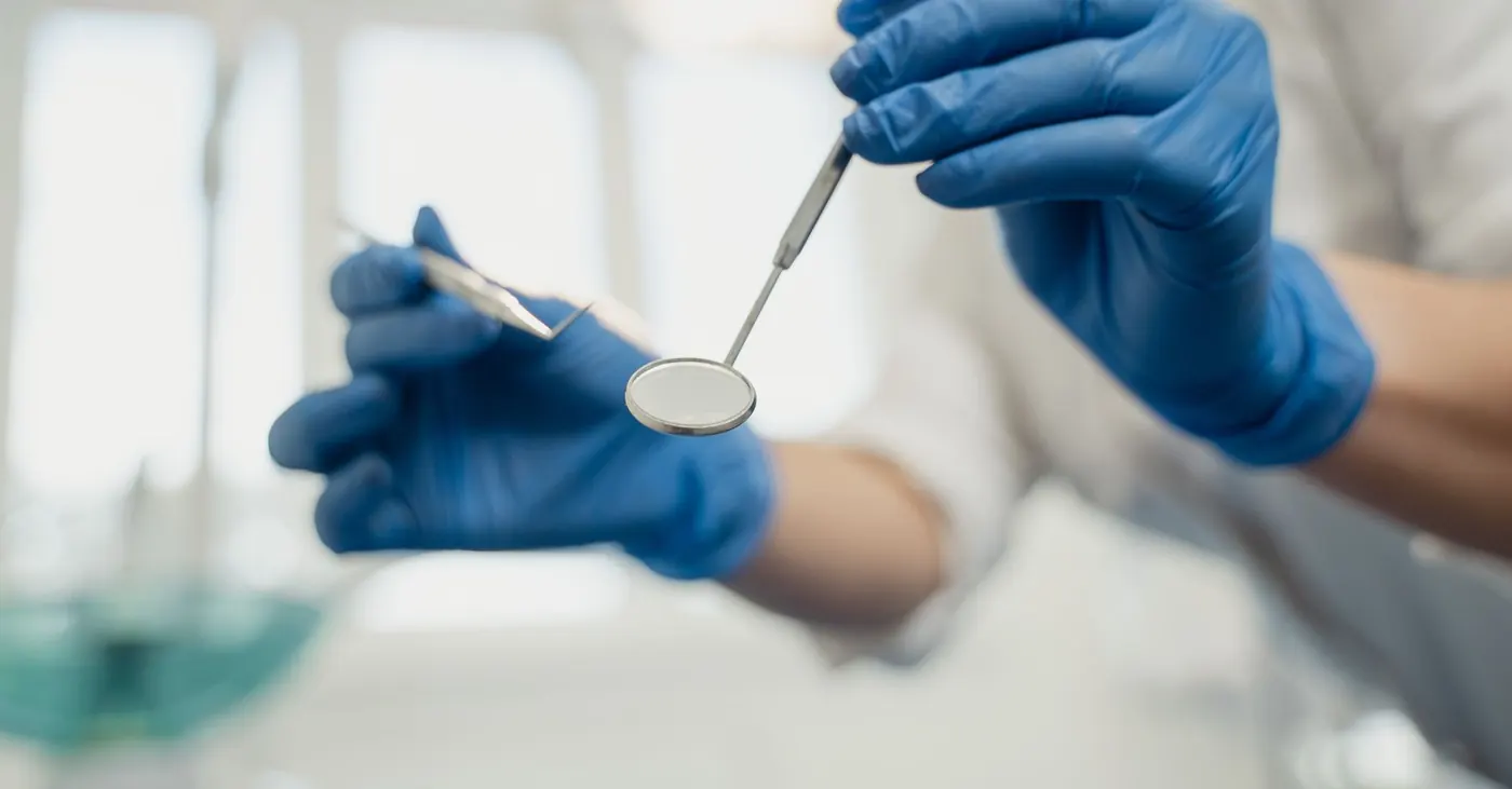 Mãos com luvas a segurar o espelho e a sonda usados na observação dos dentes